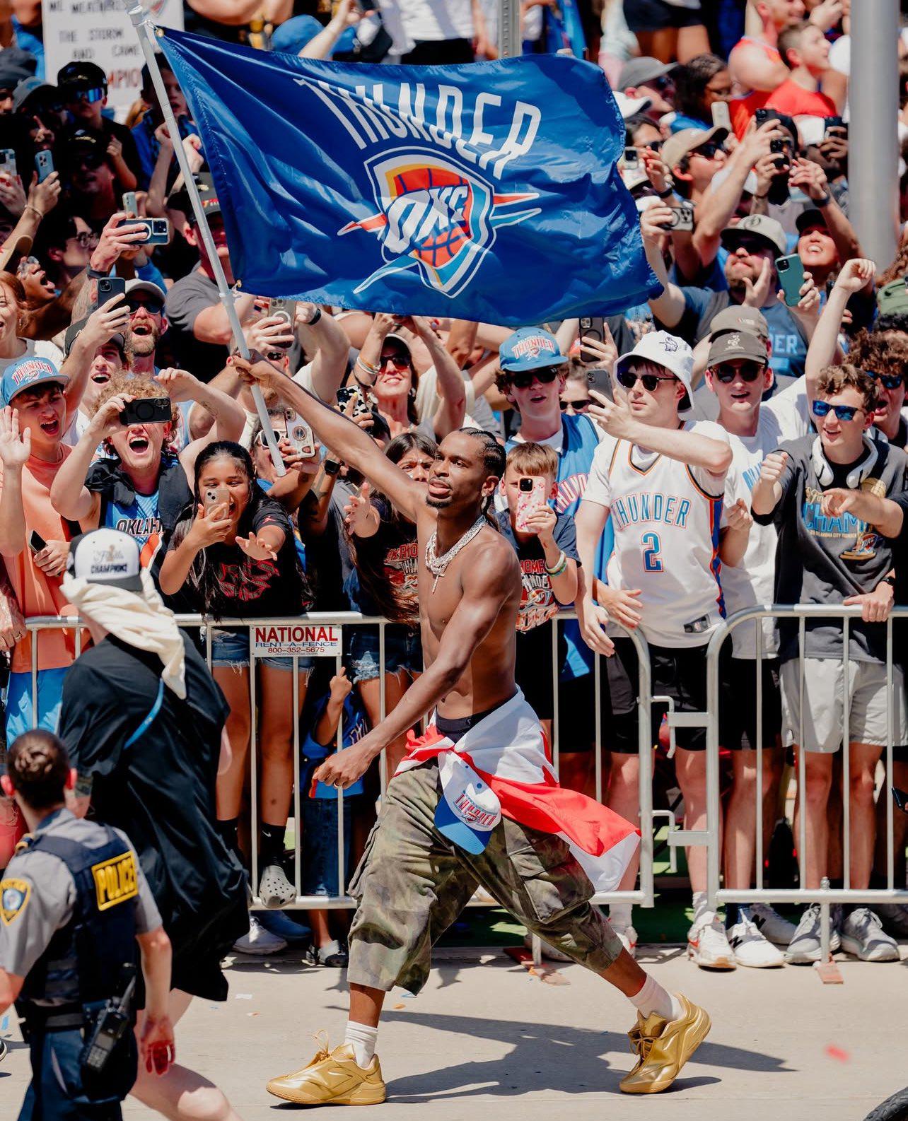 Photo of Oklahoma City Thunder superstar Shai Gilgeous Alexander, parading shirtless. He is carrying a flag with the OKC Thunder logo, and has a Canadian national flag around his waist.