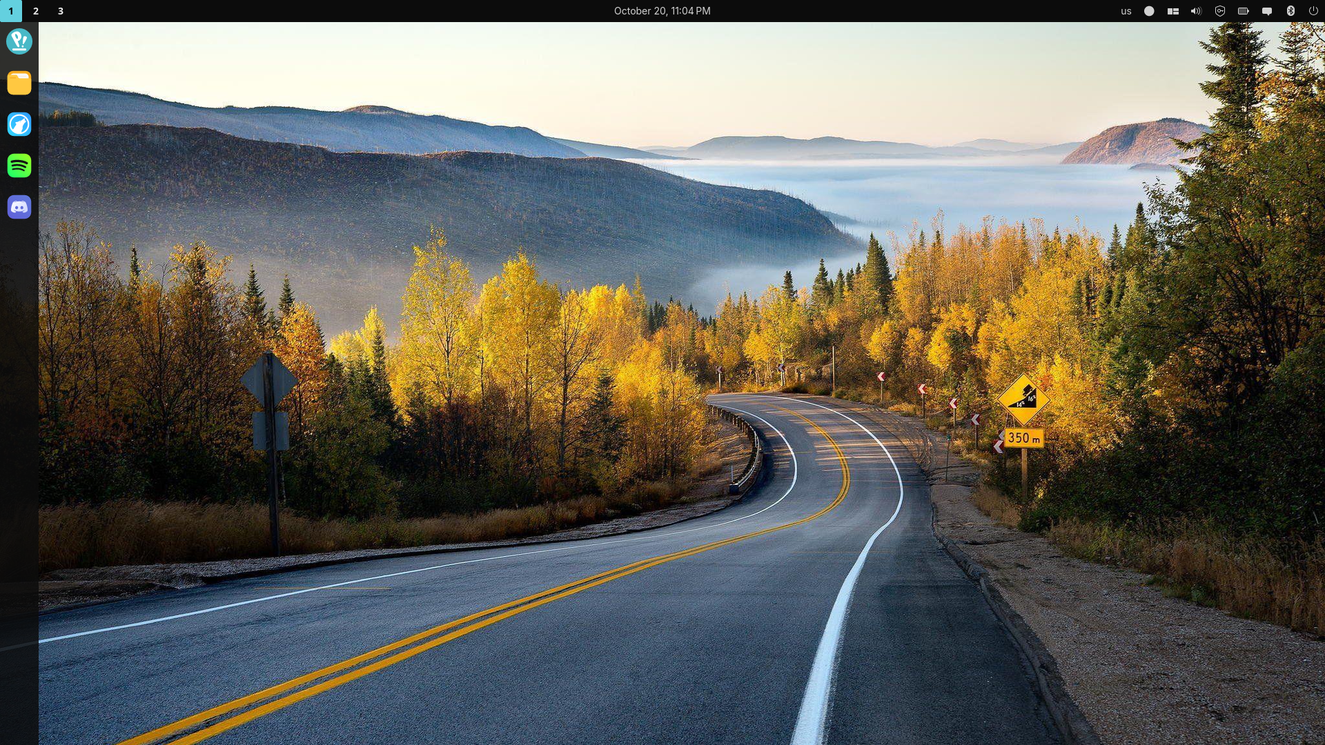 screenshot of computer desktop showcasing cosmic desktop environment. An application dock is to the left, a numbered workplace indicator is at top left, clock and calendar at top center, and system information panel at top right. Wallpaper features a mountain road in autumn.